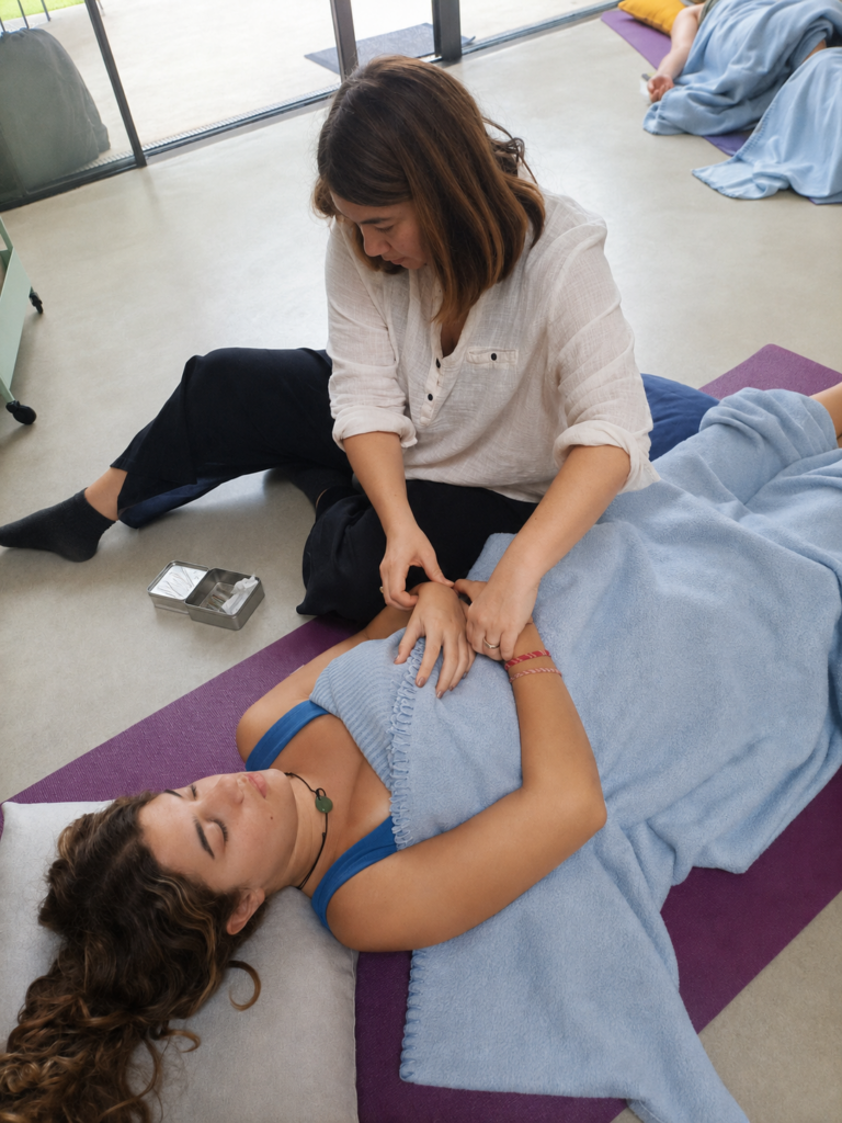Mujer tumbada sobre una esterilla durante una sesión de acupuntura comunitaria mientras la terapeuta toma el pulso